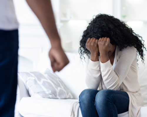 A woman on a couch covering her eyes with her hand. In front of her is a man all we can see is his closed fist.