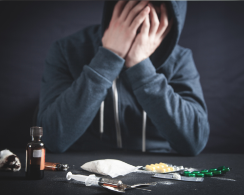 A hooded man with his hands on his face sitting at a table with lots of drugs scattered on it.