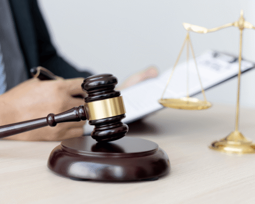 Close-up of judge’s gavel and golden scales of justice on desk, with attorney reviewing legal documents in the background, symbolizing court proceedings, bail hearings, and pretrial release decisions