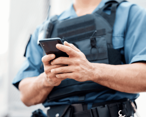 Police officer in uniform holds a smartphone while wearing a tactical vest, symbolizing police search phone without warrant LA. The image reflects cell phone search warrant California, privacy rights LA, Riley v. California application, digital searches police, LA Fourth Amendment issues, and phone search exclusions.