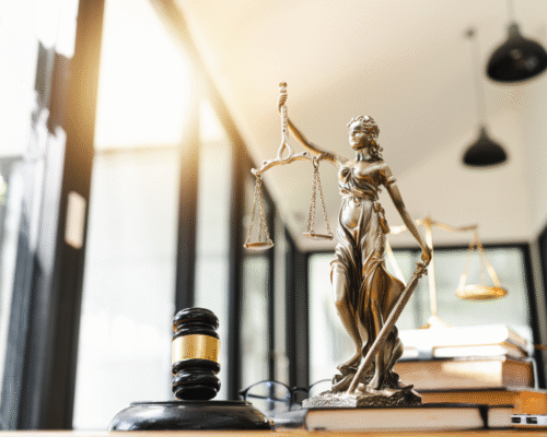 Statue of Lady Justice holding balanced scales beside a judge’s gavel and legal books on a desk in a modern office setting. The image represents the legal system and is commonly used to illustrate criminal defense topics, including cases involving revenge porn charges, consent, and intent under California law in Los Angeles.