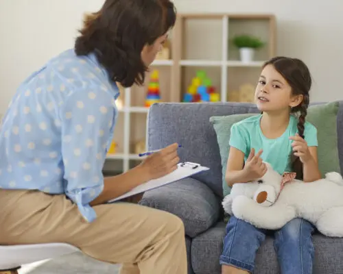 Supportive psychologist with clipboard listening to little child during therapy session. Preschool girl feeling at ease in therapist's office sharing her thoughts and concerns