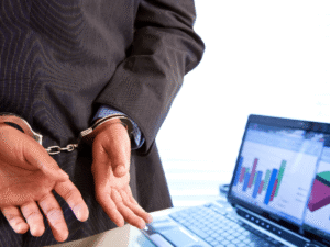 Businessperson in handcuffs near laptop displaying financial data.